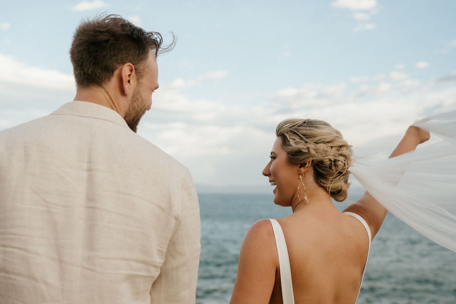 Couple in wedding attire looking at each other against ocean backdrop with flowing veil.