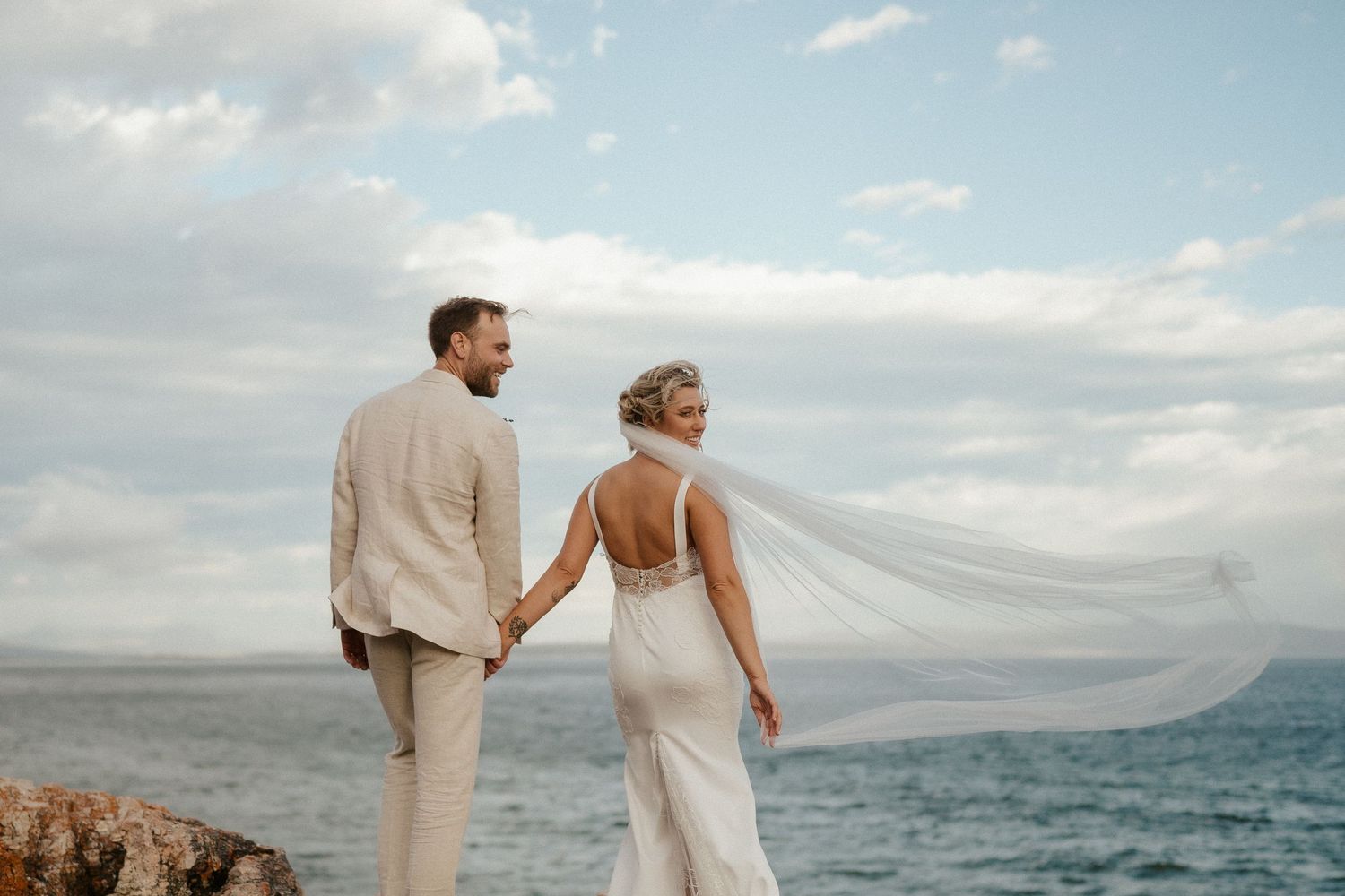 Newlyweds stand on rocky coastline with flowing veil and ocean view in background.