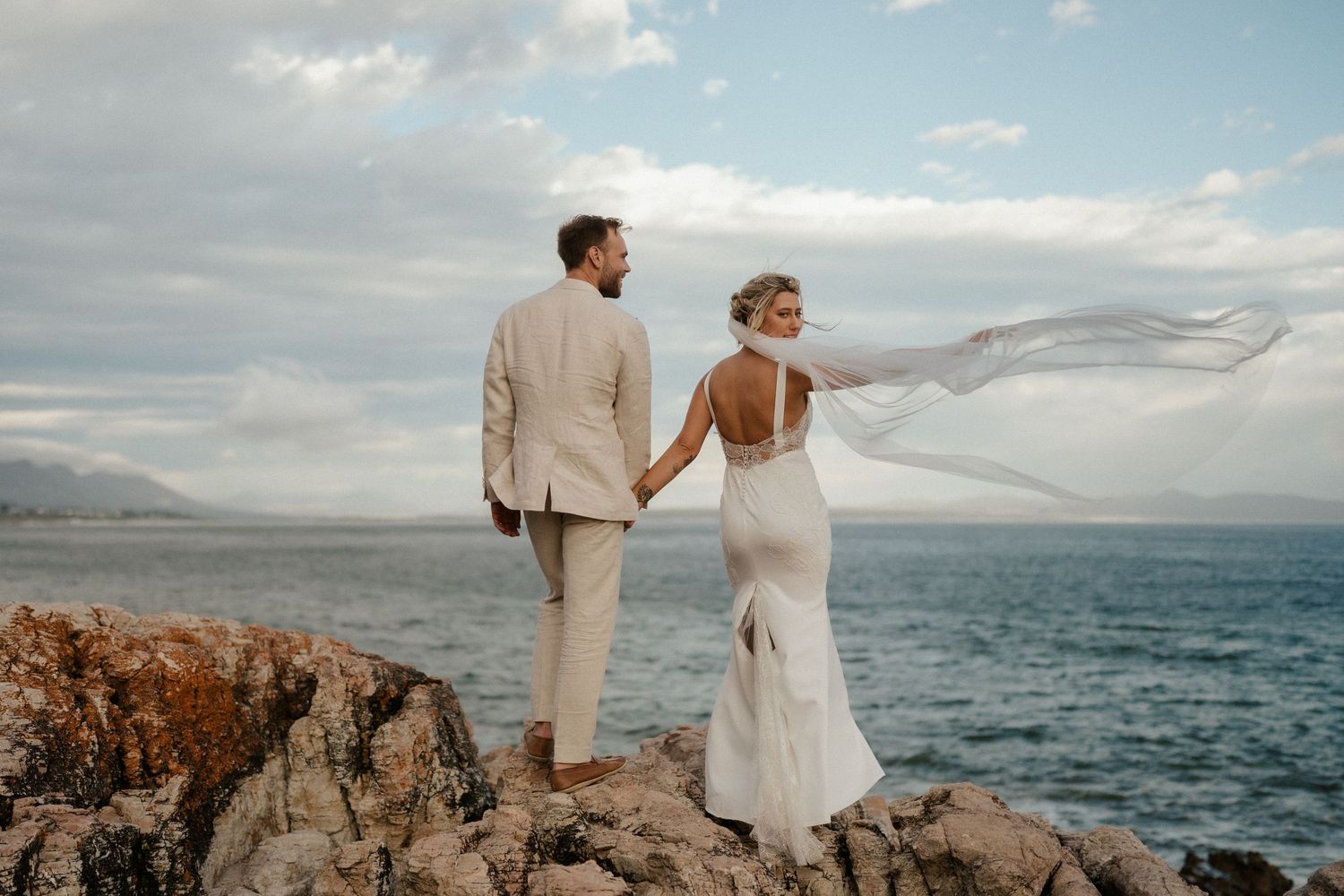 A couple stands on oceanside cliffs in elegant wedding attire as wind blows.