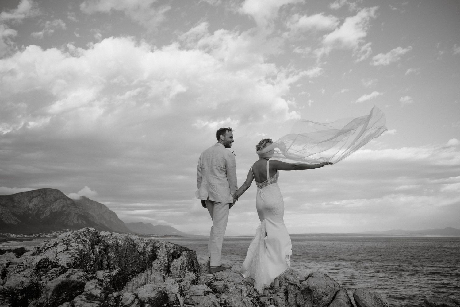 Dramatic black and white photo of a couple standing on coastal rocks with flowing veil.