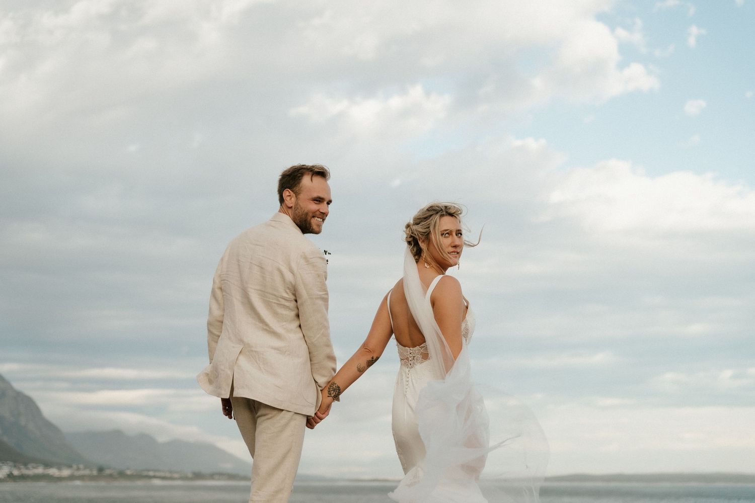 A couple walking hand in hand on a beach with cloudy skies and mountains in the background.