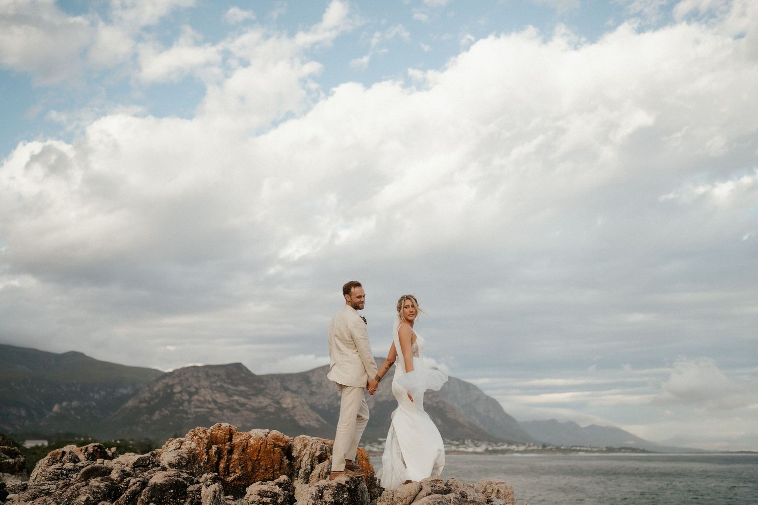 Wedding couple poses on coastal cliffs with dramatic mountain and ocean landscape.