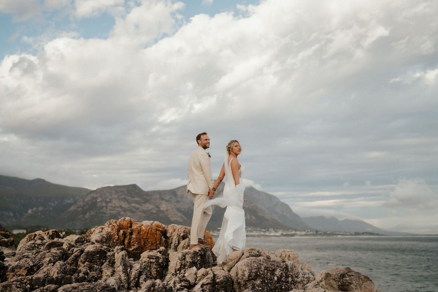 Scenic coastal wedding portrait with couple standing on rocks overlooking mountain bay.