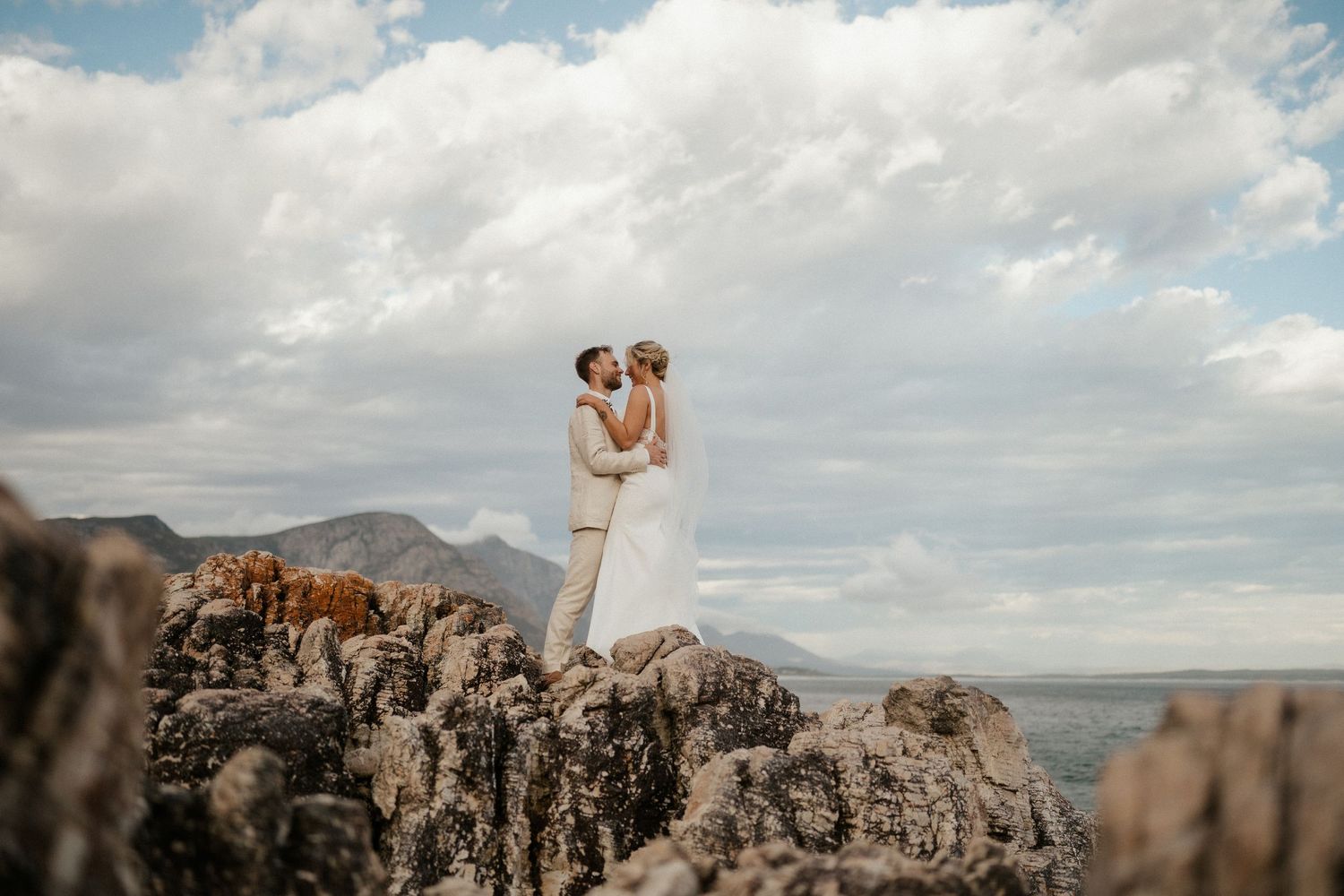 Newlyweds embrace on dramatic rocky cliffs with mountains and cloudy skies in background.