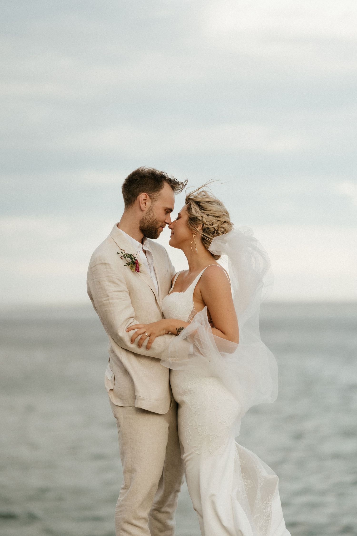 A couple in light-colored wedding attire embrace by the ocean on their special day.