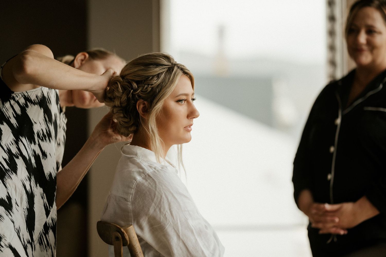 Wedding preparations in progress as a stylist works on arranging the bride's hair in a sunlit room.