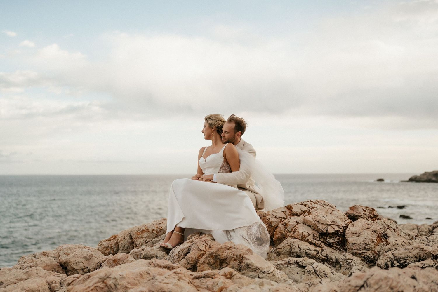 Couple in wedding attire sitting together on coastal rocks overlooking the ocean at sunset.