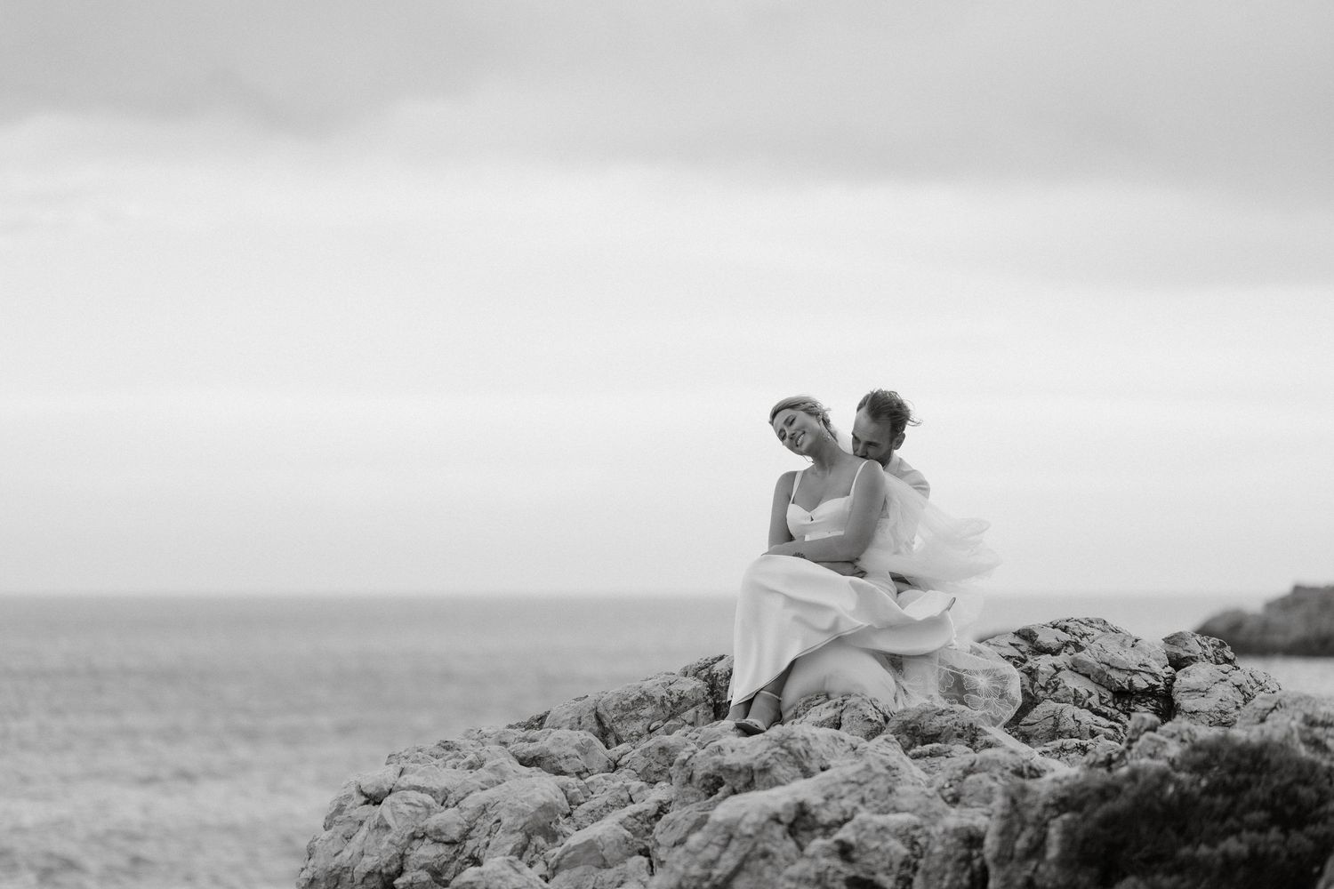 A black and white photo shows a couple embracing on coastal rocks overlooking the ocean.