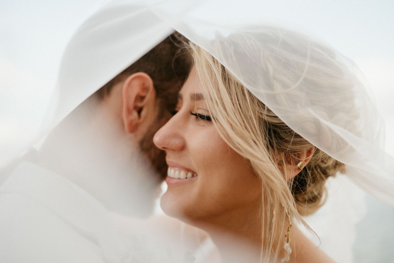 A tender moment shared under a flowing white veil against a bright sky.