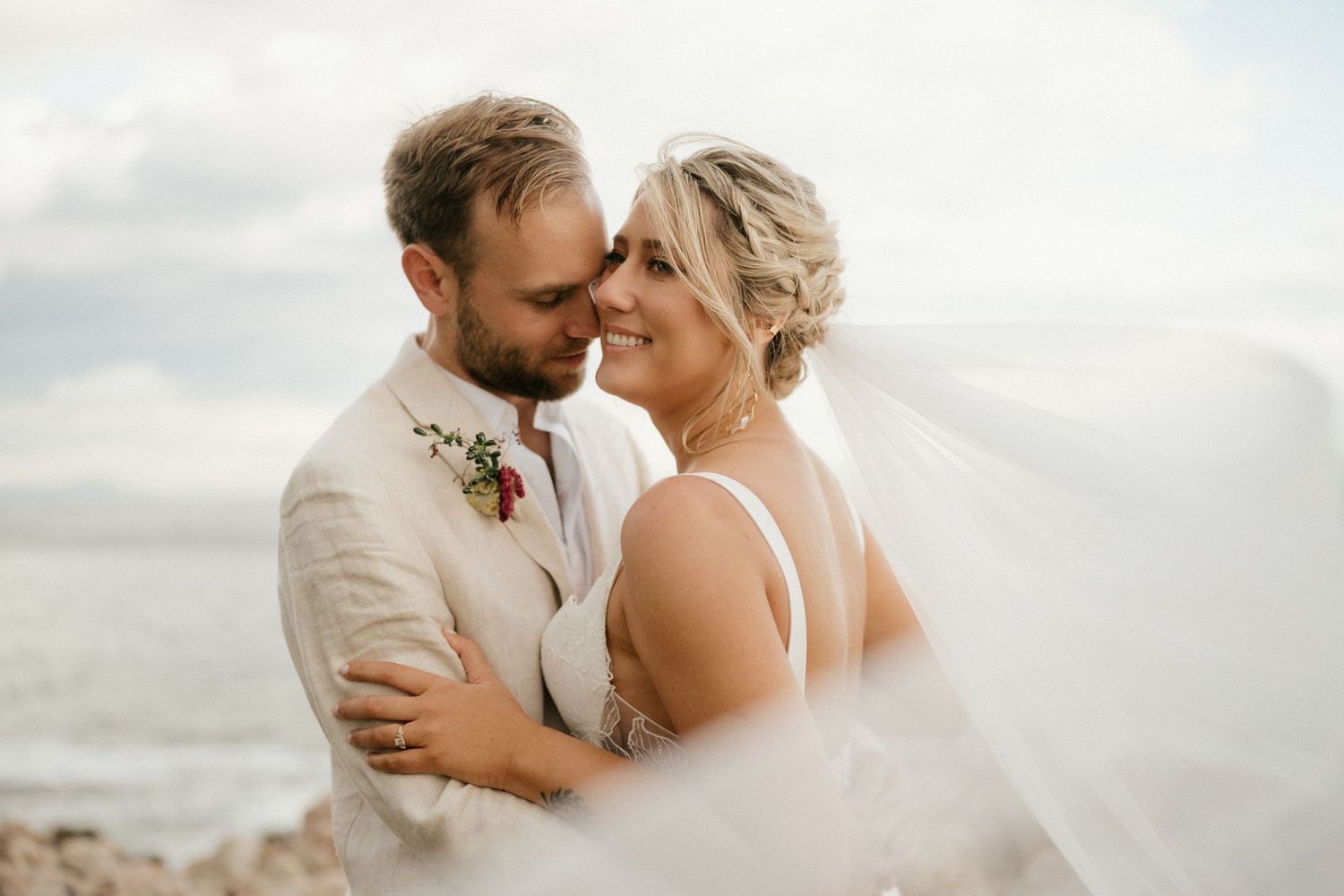 An intimate moment between newlyweds as a veil flows in the coastal breeze.
