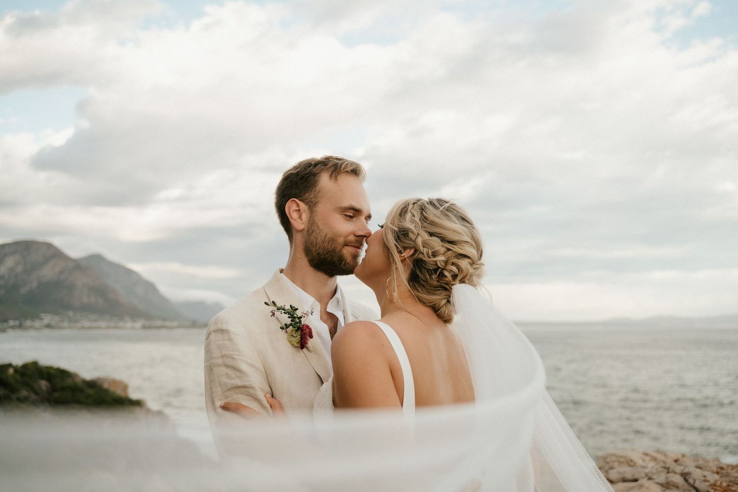 A wedding couple embraces on a scenic coastal overlook with mountains in the background.