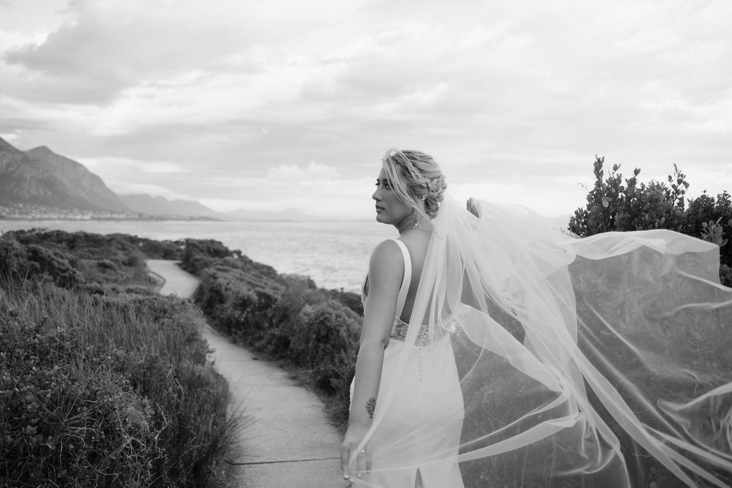 A bride with flowing veil walks along coastal path with mountains in background.