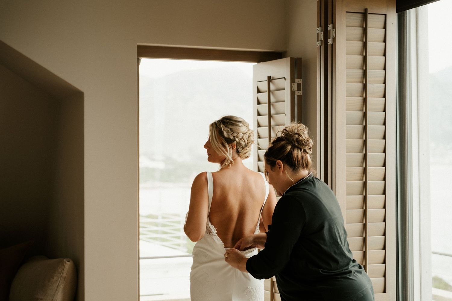 A wedding dress being fastened near a window with bright natural light streaming in.