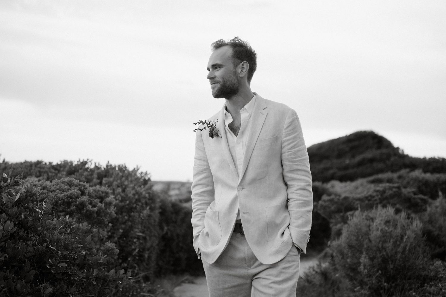 A well-dressed man in light suit stands thoughtfully against coastal landscape.