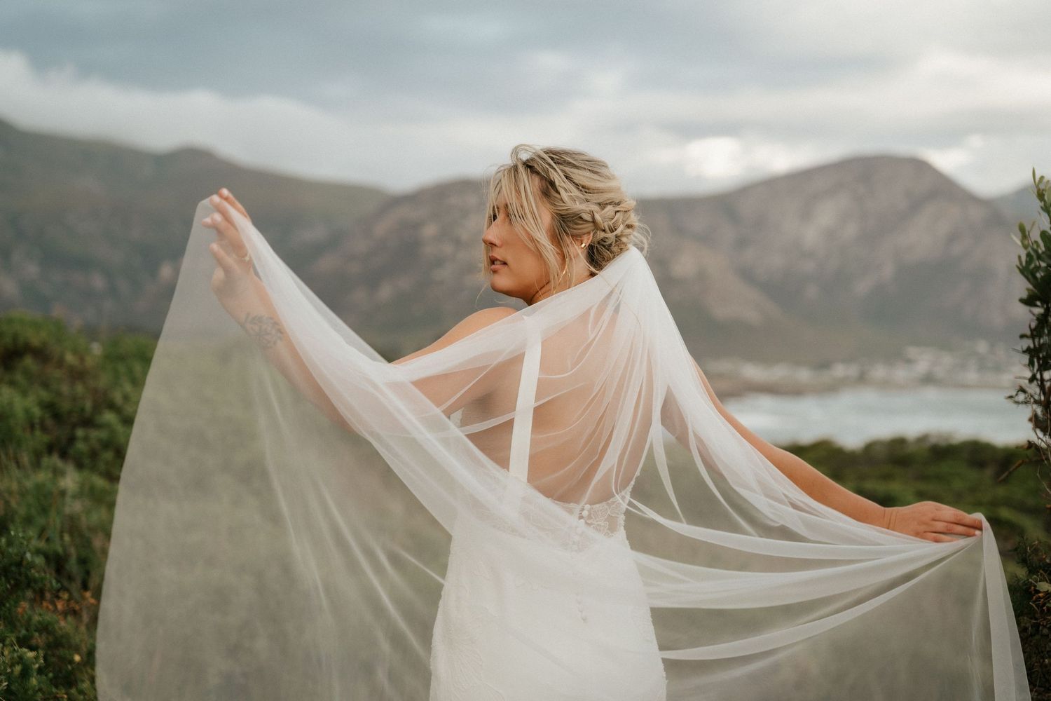 A bride in a white dress holds her veil flowing in the wind against mountain scenery.