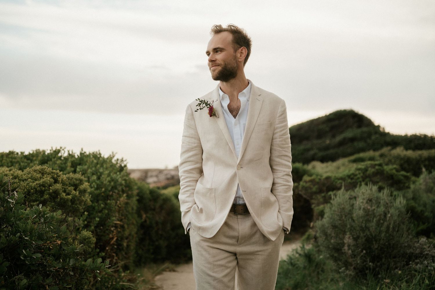 A groom in a light colored suit stands against a natural landscape at sunset.