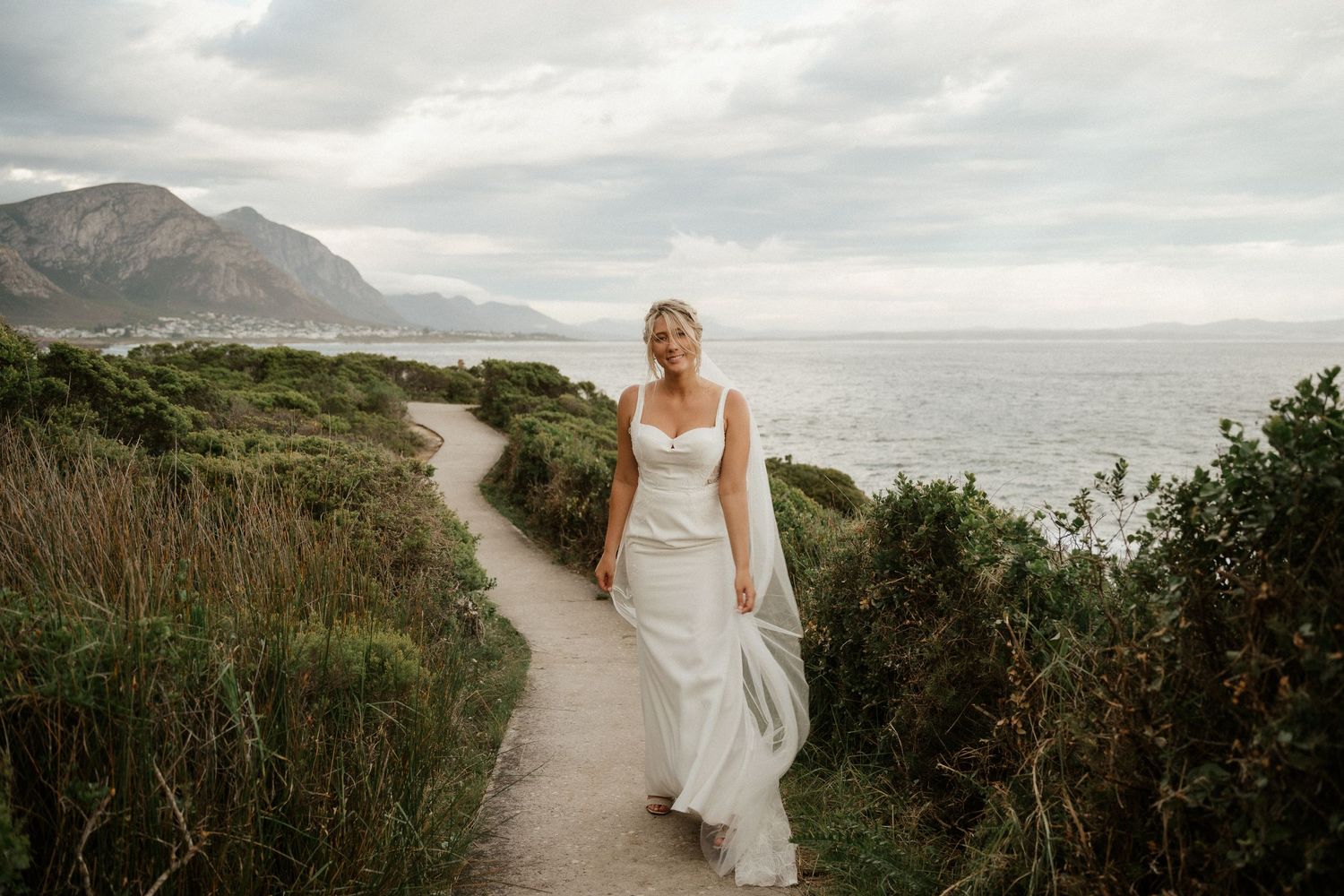 White wedding dress flowing in the coastal breeze along a scenic mountain path by the ocean.