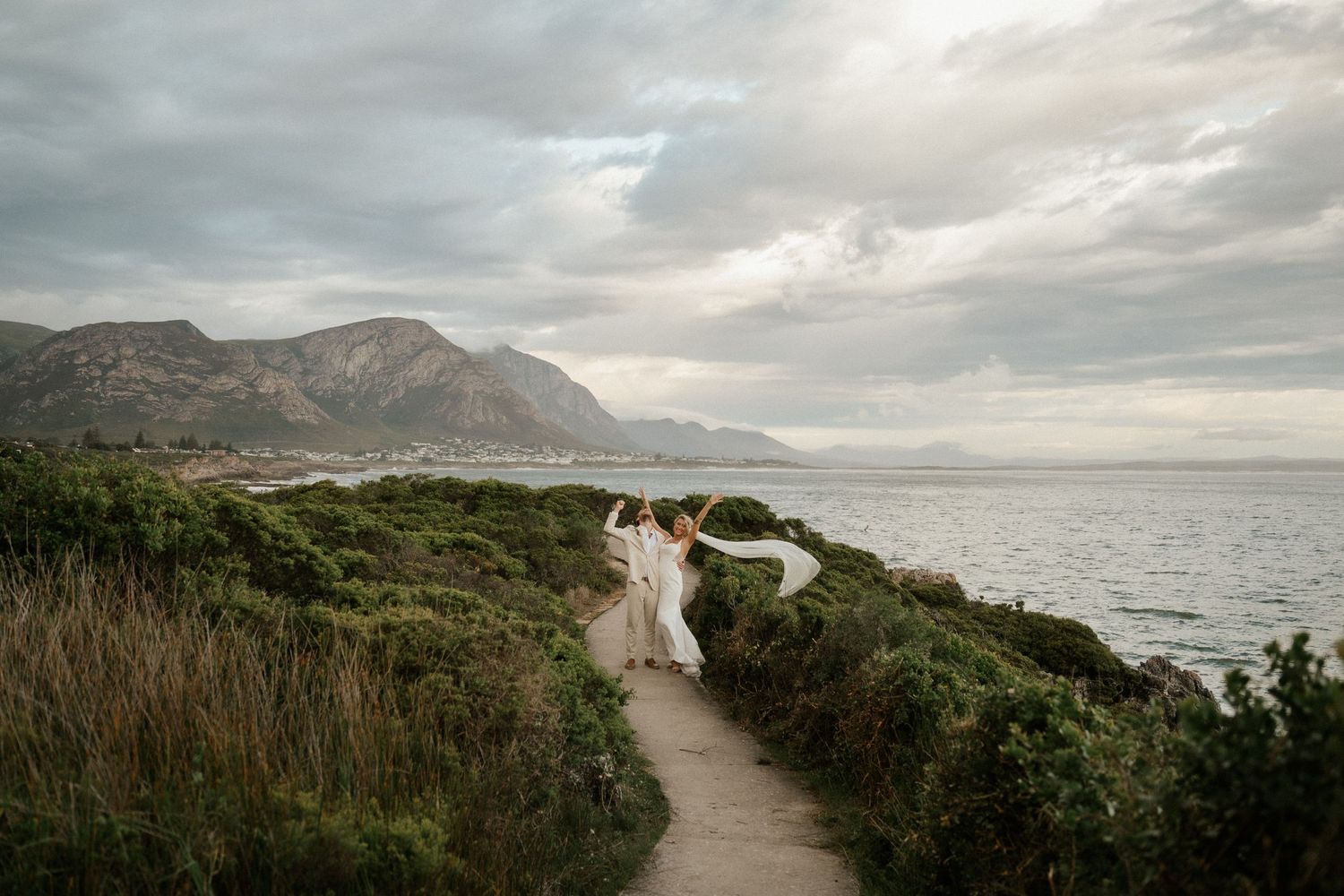 A couple in wedding attire stands on a coastal path with dramatic mountain cliffs and ocean views in the background.