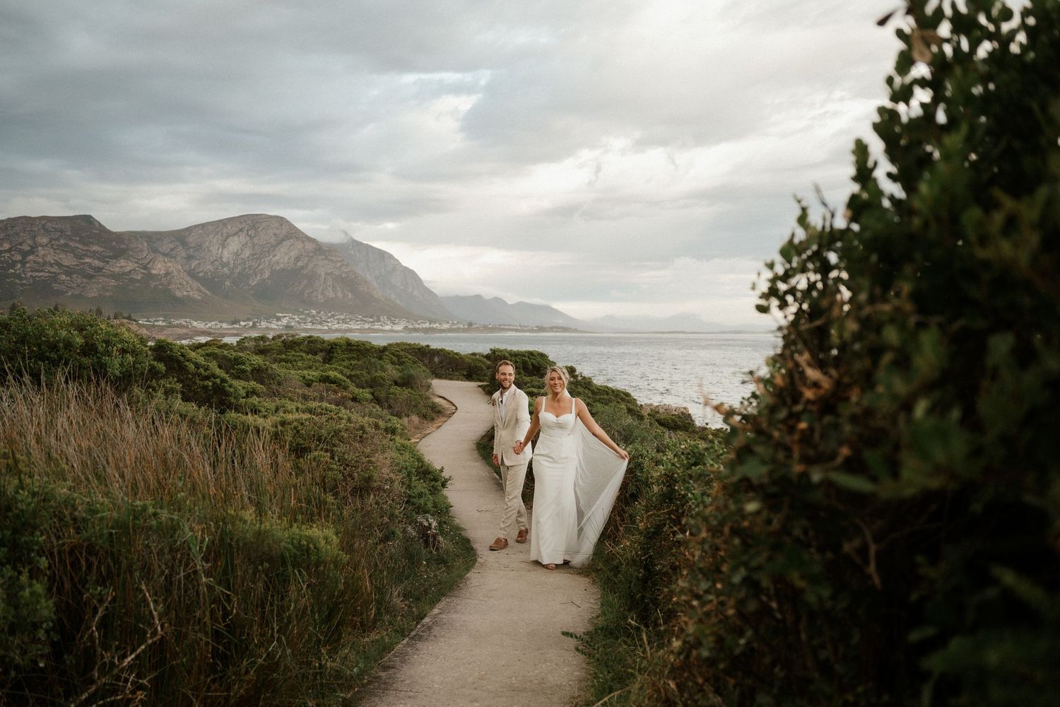 A couple walks down a coastal path surrounded by lush vegetation with mountains in the background.