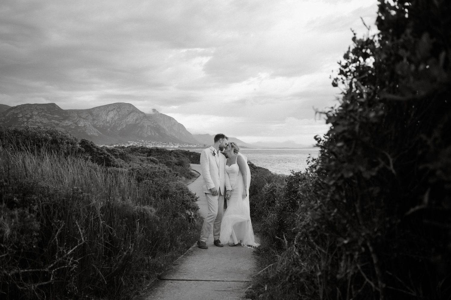 Black and white photo of a couple walking along a coastal path with mountains in the background.