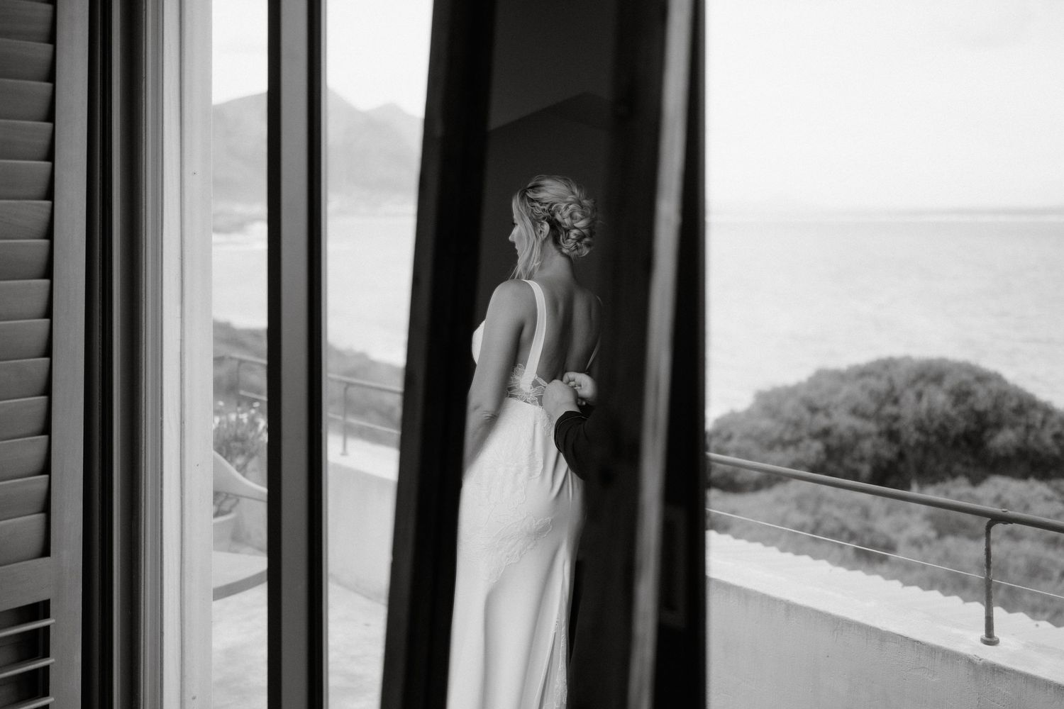 A silhouetted figure in an elegant white dress stands by a window overlooking an ocean view.