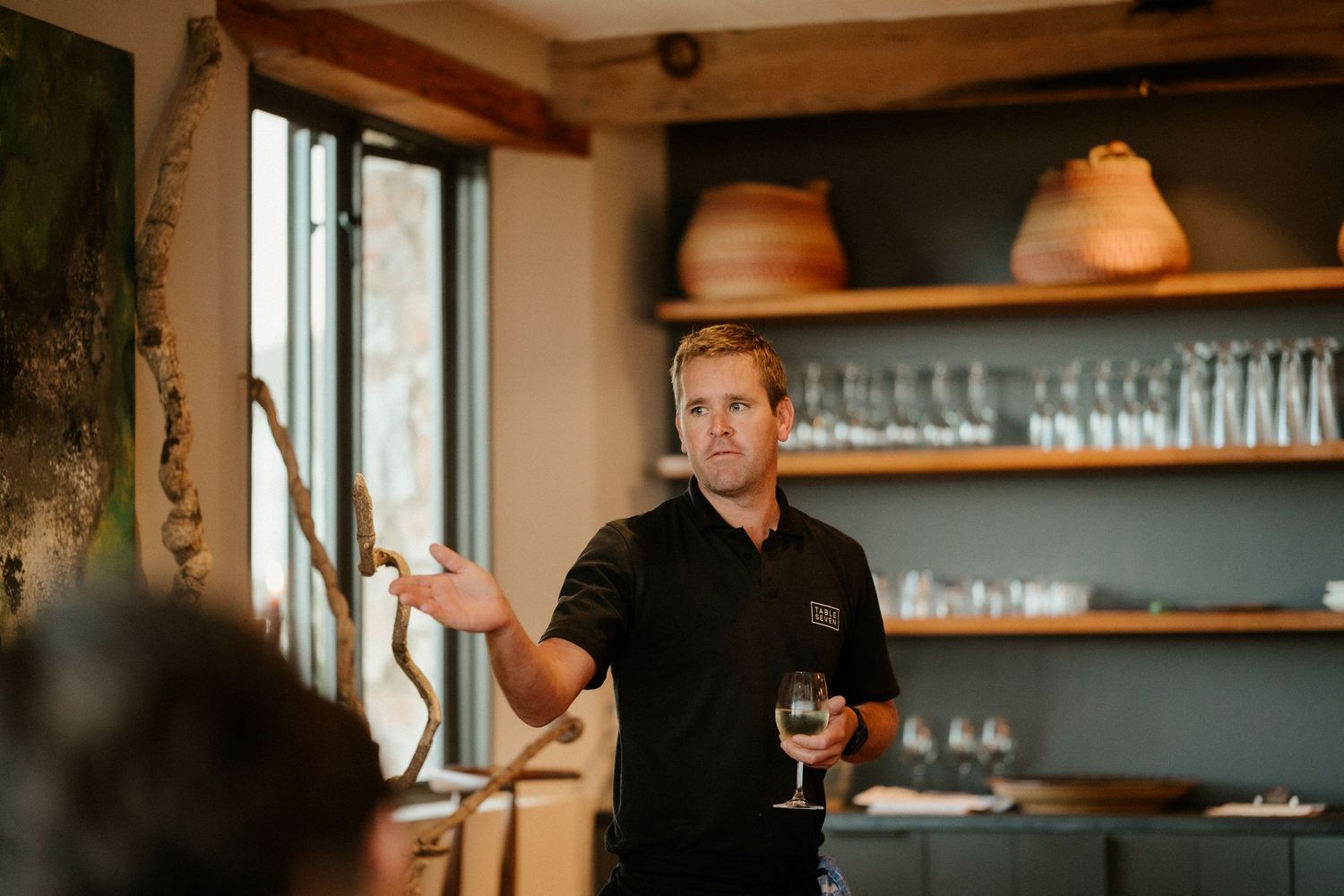 Restaurant staff member speaking with wine glass in hand at cozy establishment with shelving and glassware.