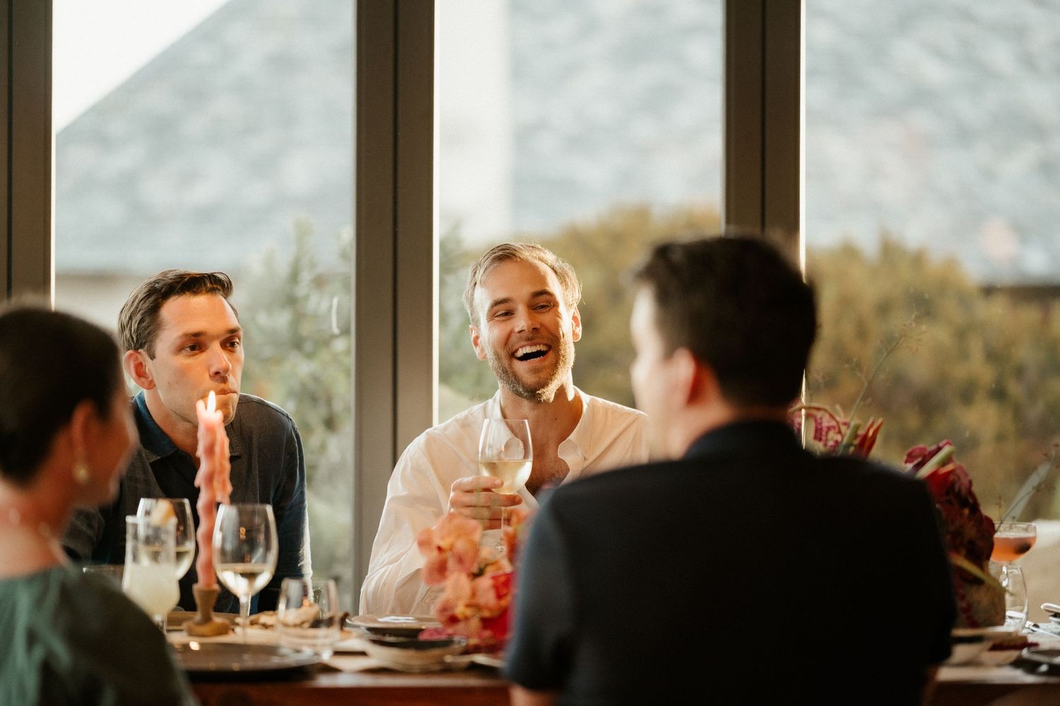 A lively dinner party scene with guests enjoying wine and conversation at a table with mountain views in the background.