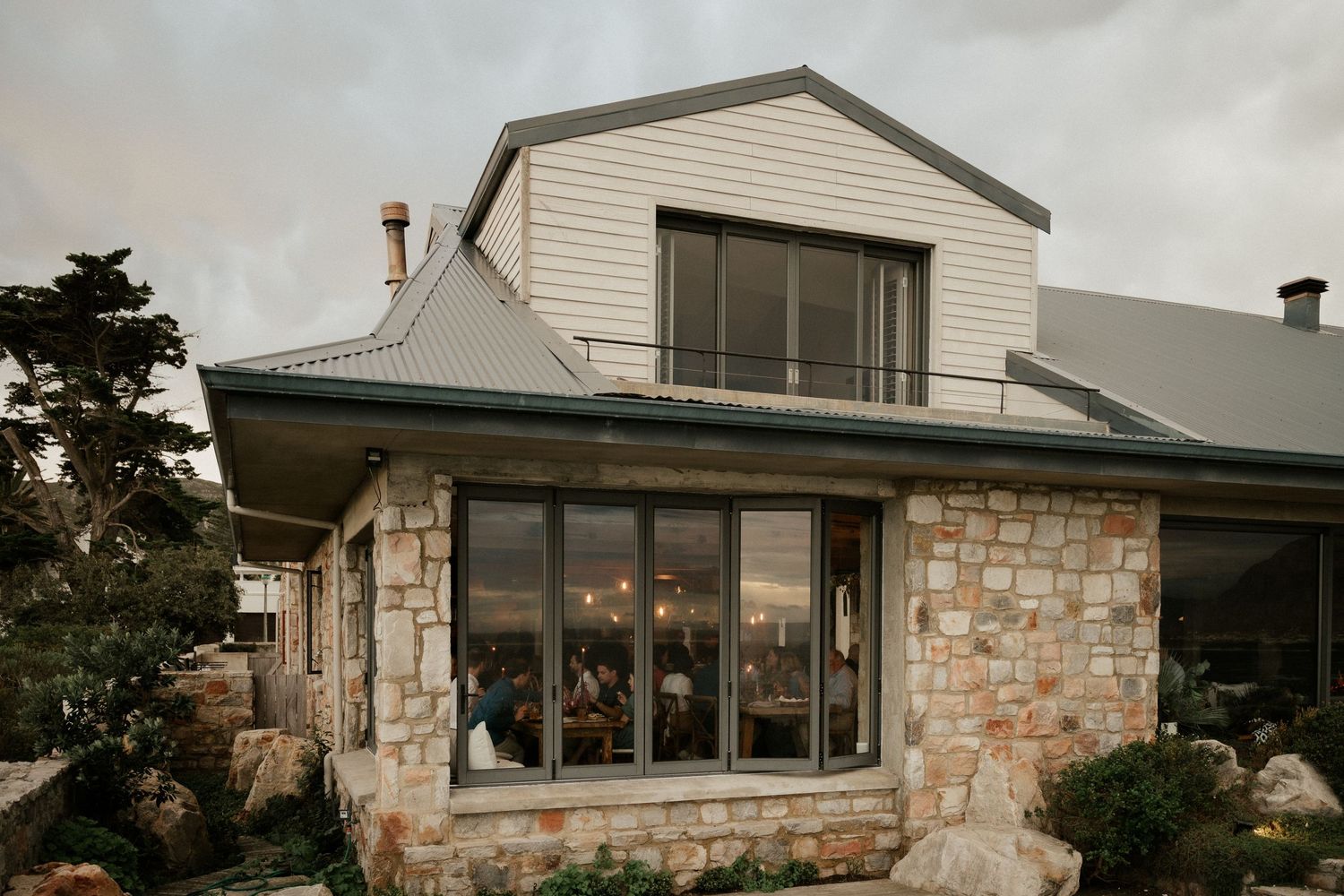 Modern rustic house exterior with stone facade and large windows reflecting evening light.