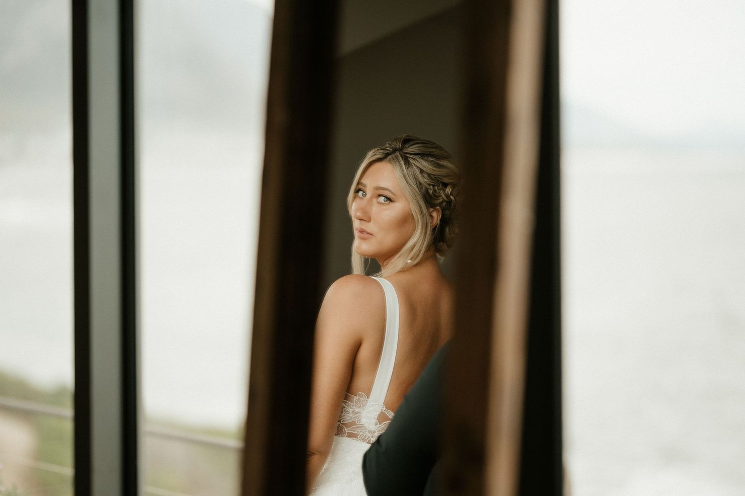 A bride in a sleek white dress stands by a window in a dreamy profile shot during wedding preparations.