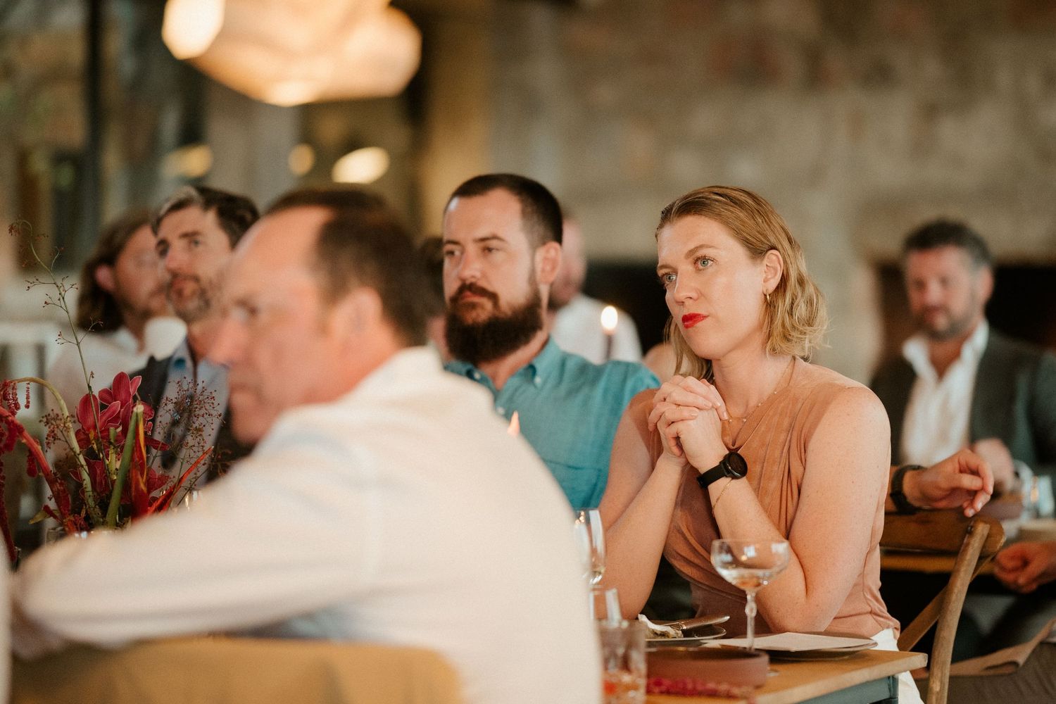 Guests attentively listen during an indoor dining event with red floral decorations on the tables.