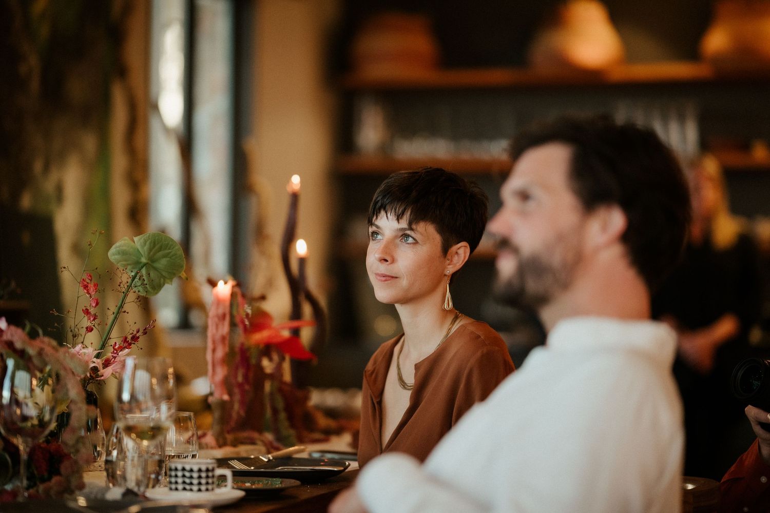 Candlelit dinner party with guests seated at an elegantly decorated table with greenery and warm lighting.