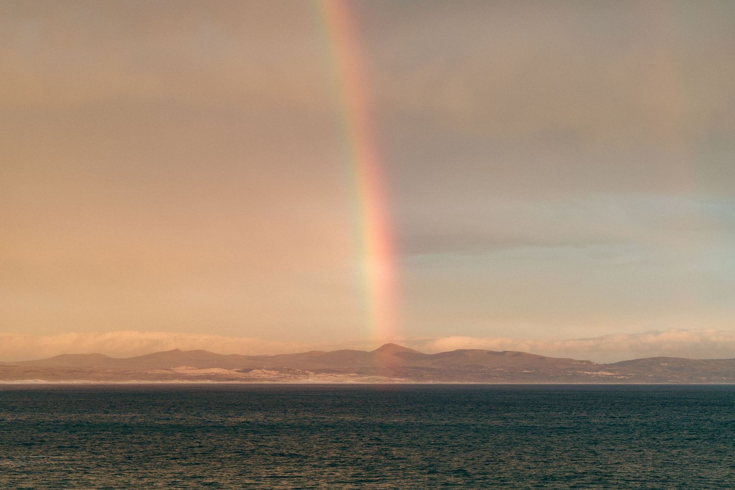 A vibrant rainbow appears over mountains and ocean during golden sunset.