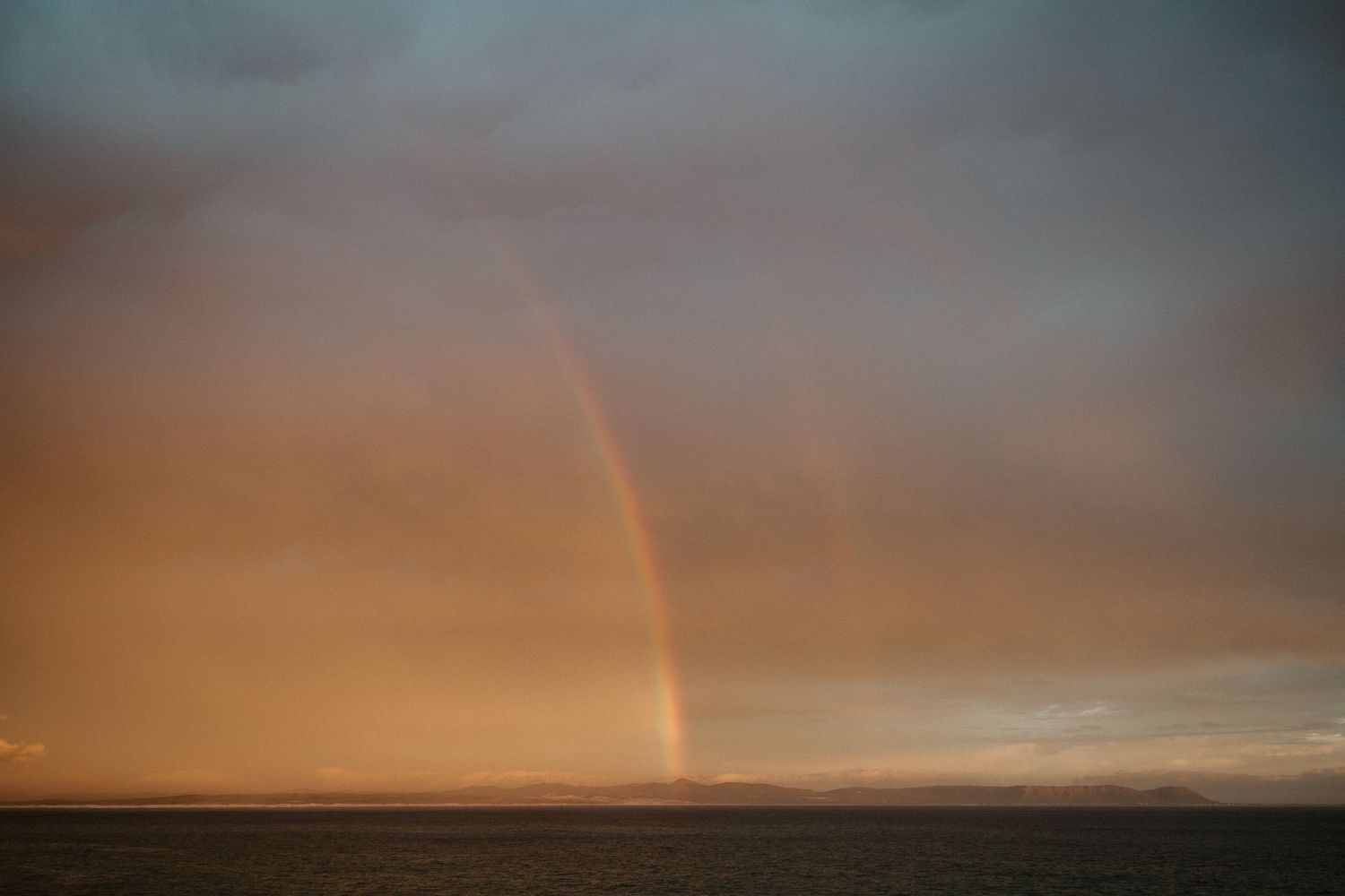 Vibrant rainbow appears in golden sunset sky over distant horizon.