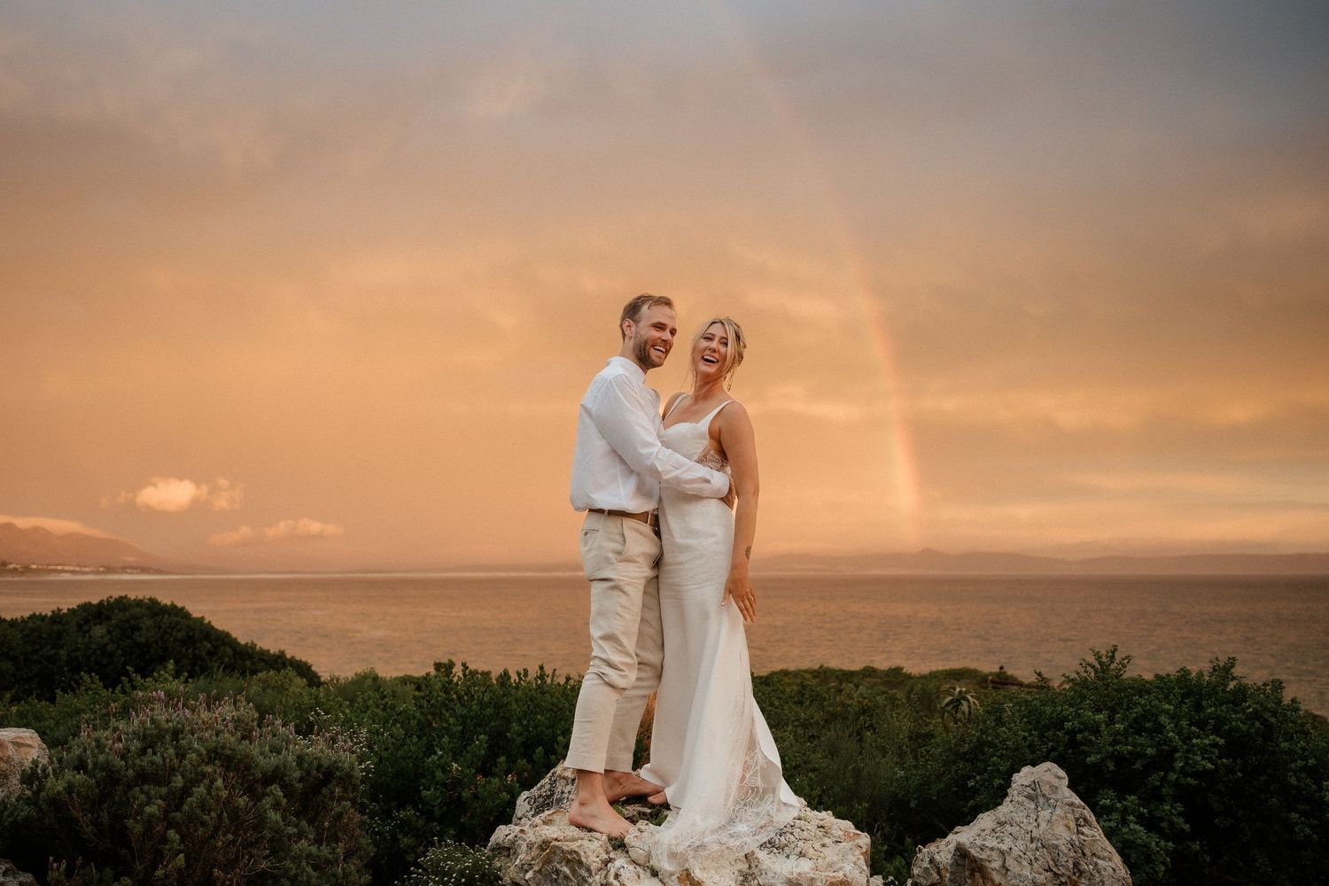 Newlywed couple stand on rocky cliff overlooking ocean during golden sunset.