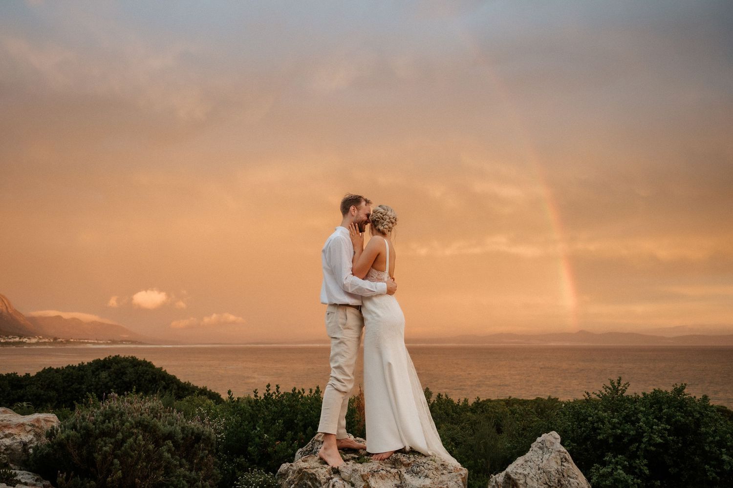Sunset wedding portrait on rocky overlook with golden sky and rainbow in background.