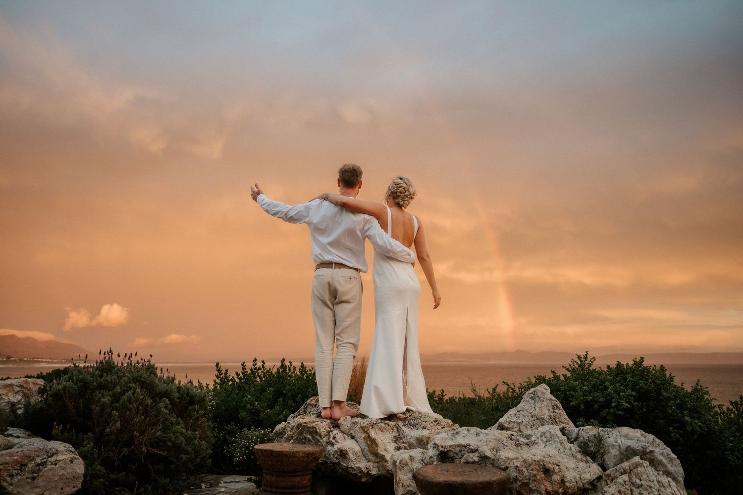 Couple stands triumphantly on rocky overlook during golden sunset.