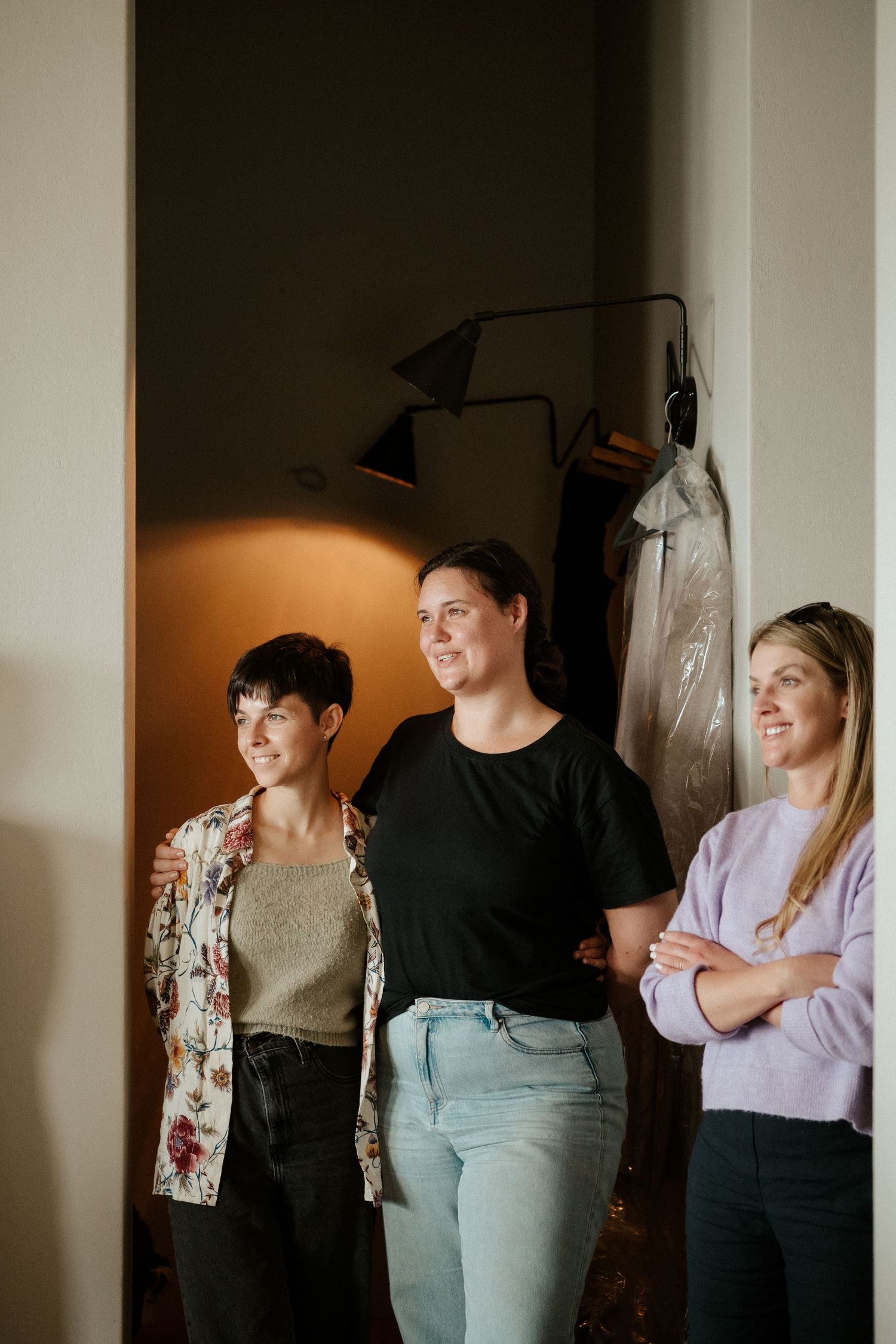 Three people standing together in casual attire smile while looking off to the side in an interior setting.