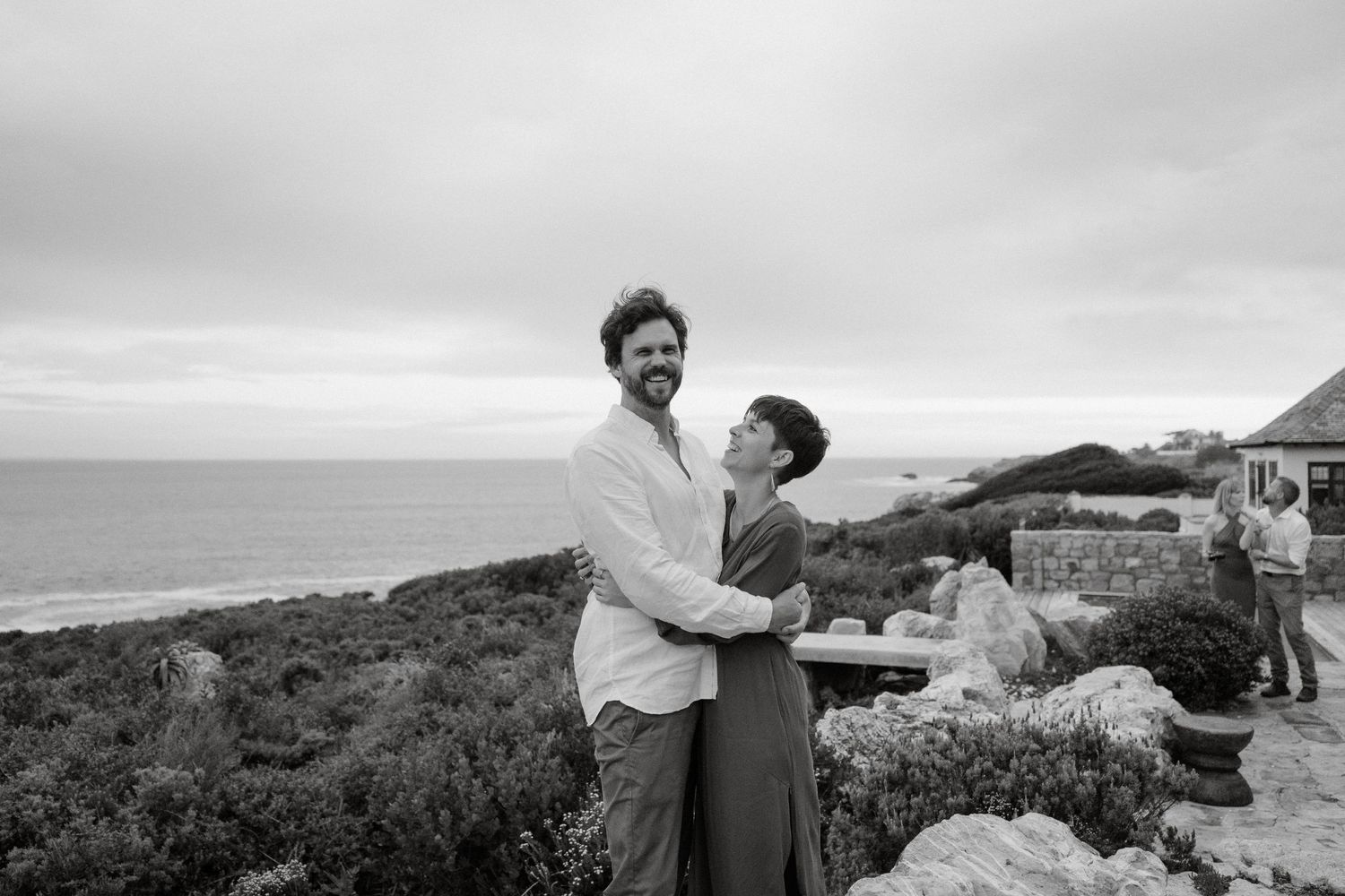 A couple embraces while standing on a coastal cliff with an ocean view in the background.