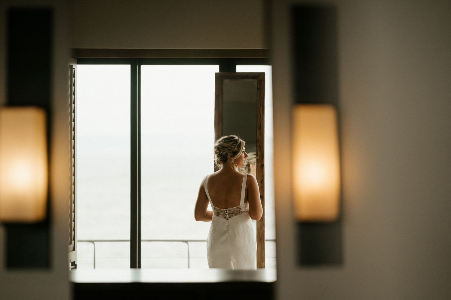 A silhouette of someone in a white dress standing by a window with warm lighting.