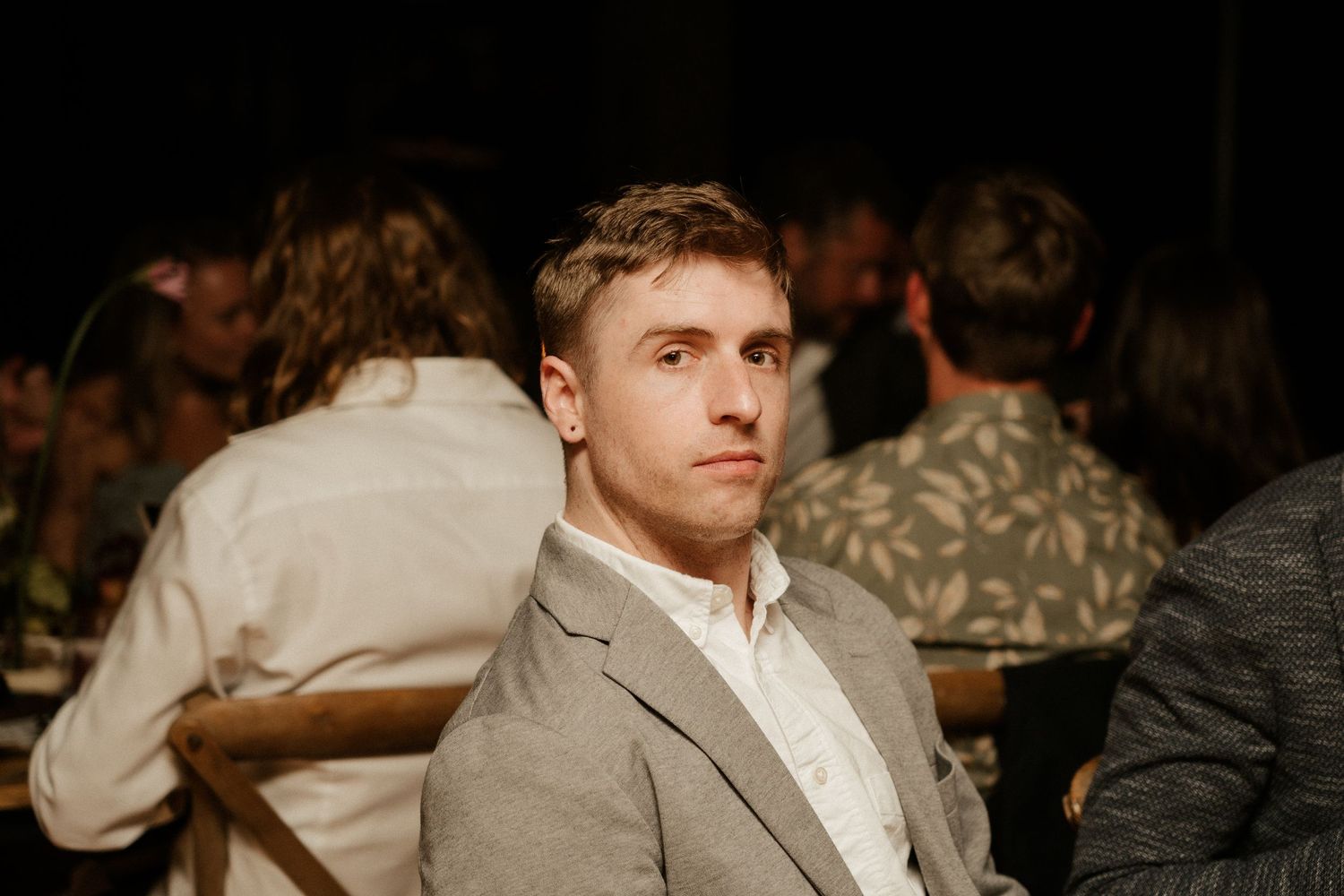 A guest in a light gray suit jacket sits thoughtfully at a dimly lit indoor event.