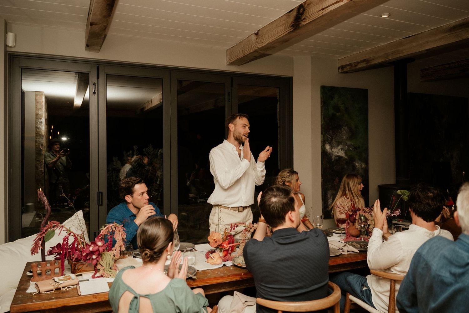 Wedding reception dinner with someone giving a speech to attentive guests at a long table.