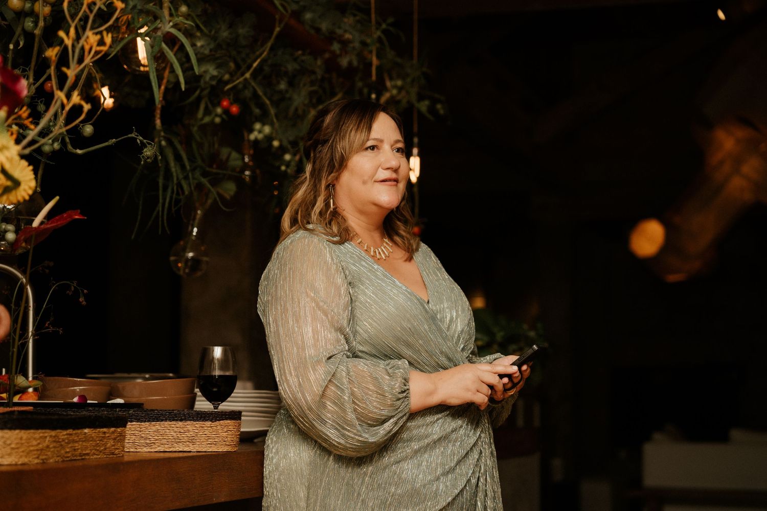 A guest at an evening reception stands in front of a floral display wearing a beige lace dress with wine nearby.