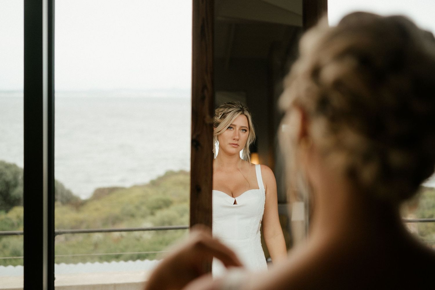 Bride looking in mirror with ocean view visible through window.