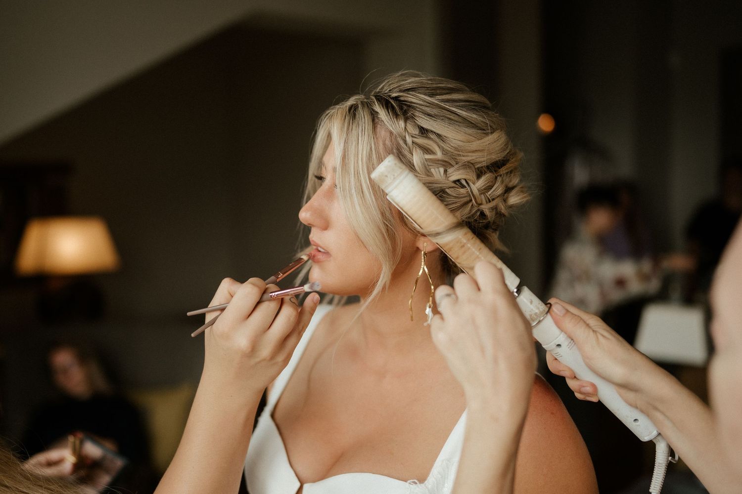 Final touch ups and makeup being applied during bridal preparation with warm lighting in a dimly lit room.