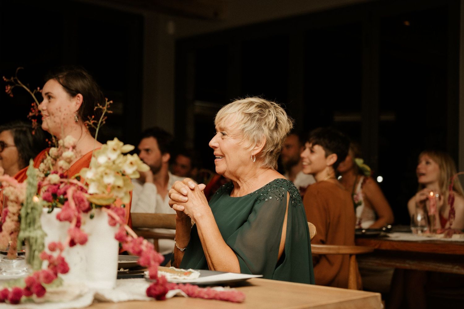 Wedding guest in a dark green dress claps during reception celebration with pink floral centerpieces on table.