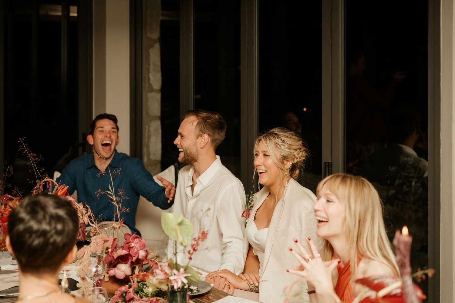 Wedding reception guests laughing together at a decorated dinner table with floral arrangements.