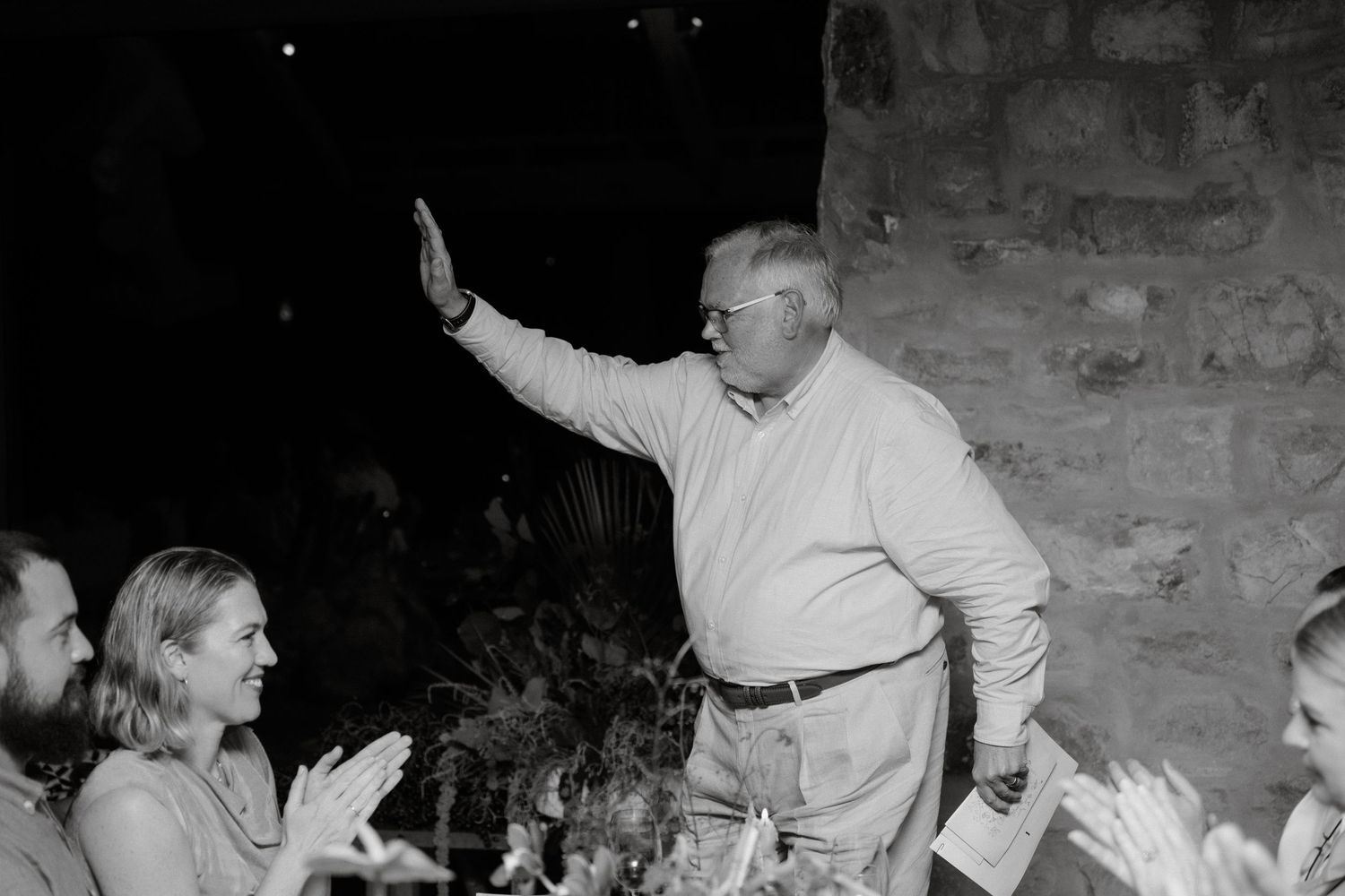 A speaker raises his hand while addressing an engaged audience in a rustic stone venue.