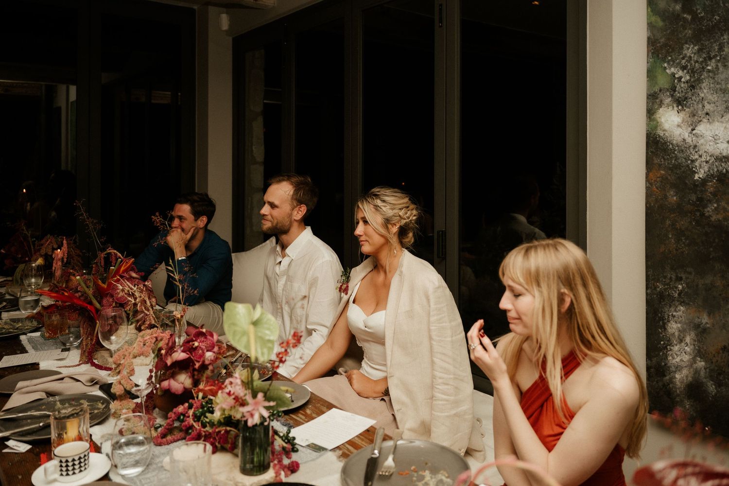 Wedding reception dinner table with guests and vibrant red floral centerpieces.