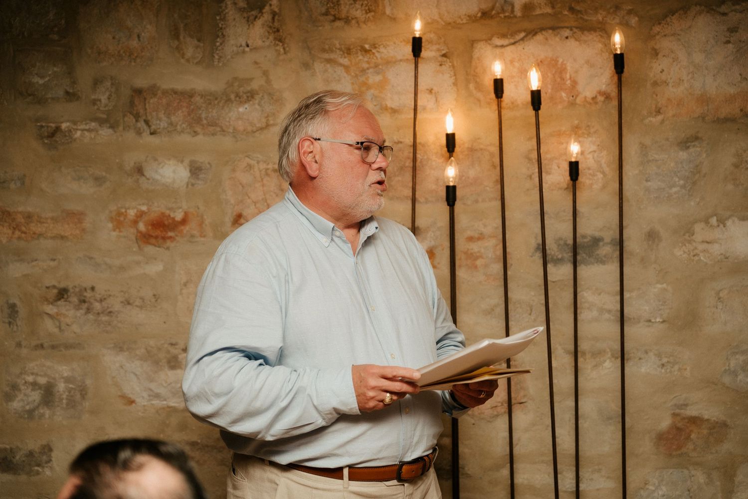 Person giving a speech in front of candlelit stone wall during evening celebration.