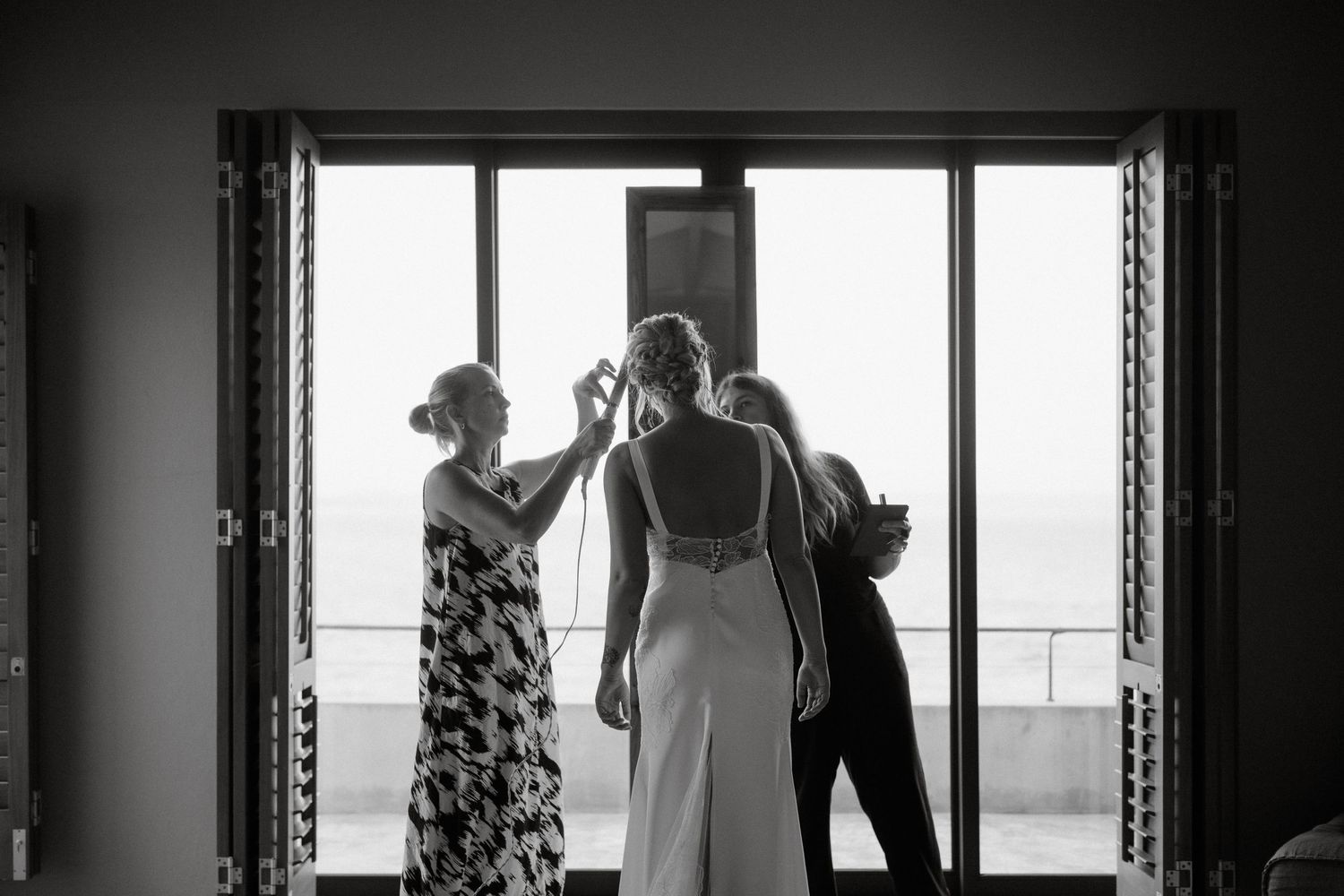 Wedding preparations take place in front of large windows as attendants assist with final details.